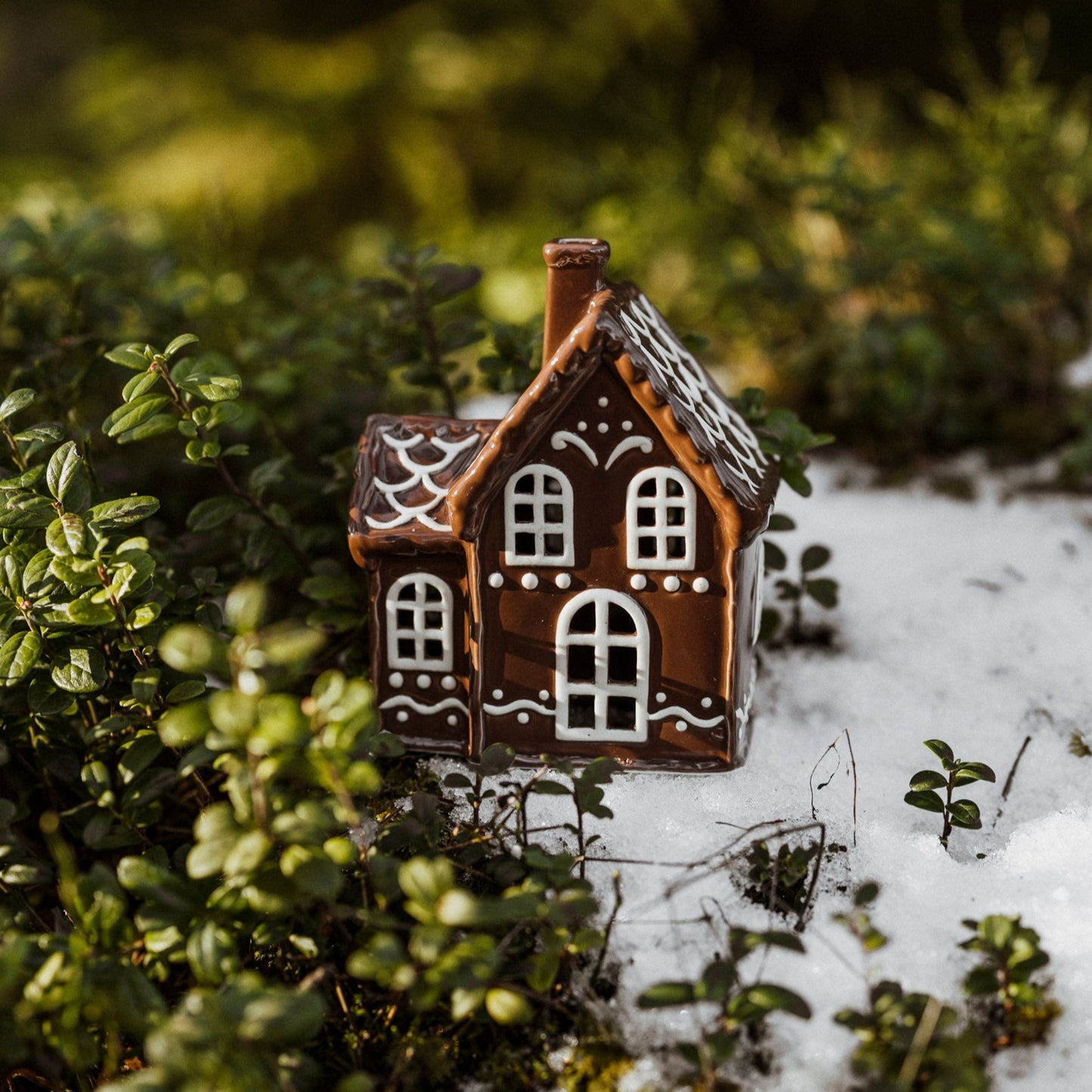 A small ceramic house with a gingerbread design placed on a bed of snow and greenery, resembling Christmas decorations.