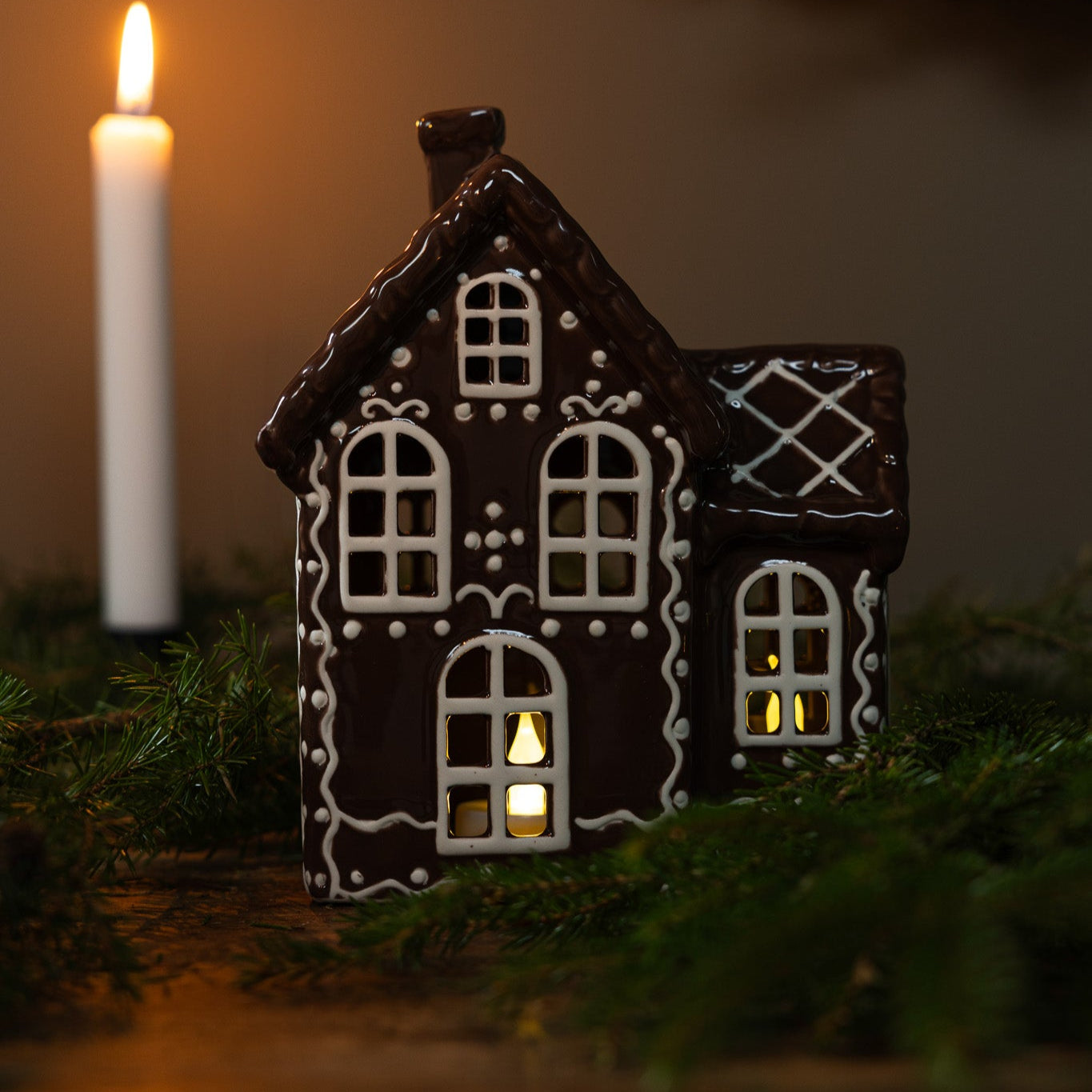 A ceramic house with a gingerbread design, decorated in black and white, sitting on a bed of greenery with a lit candle beside it, creating a Christmas decoration theme.