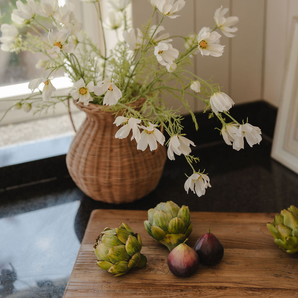 Vase with white flowers on a wooden table next to artichokes and plums.