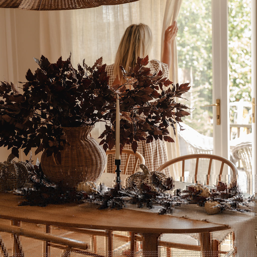 Dining room with a table set for a meal, featuring a vase of dried plants and a person in the background.