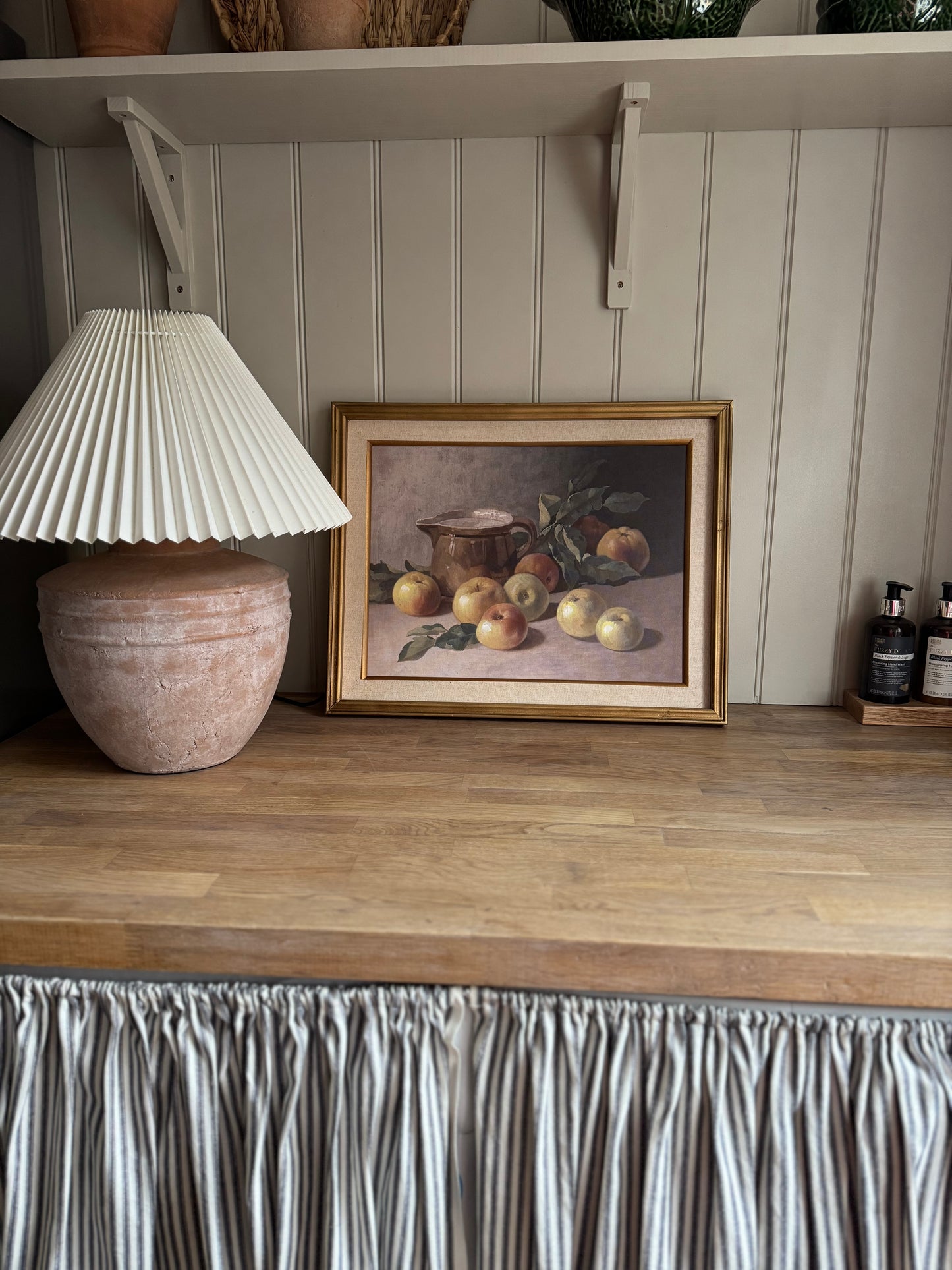 Framed still life painting of apples on a wooden shelf with a lamp and decorative items.