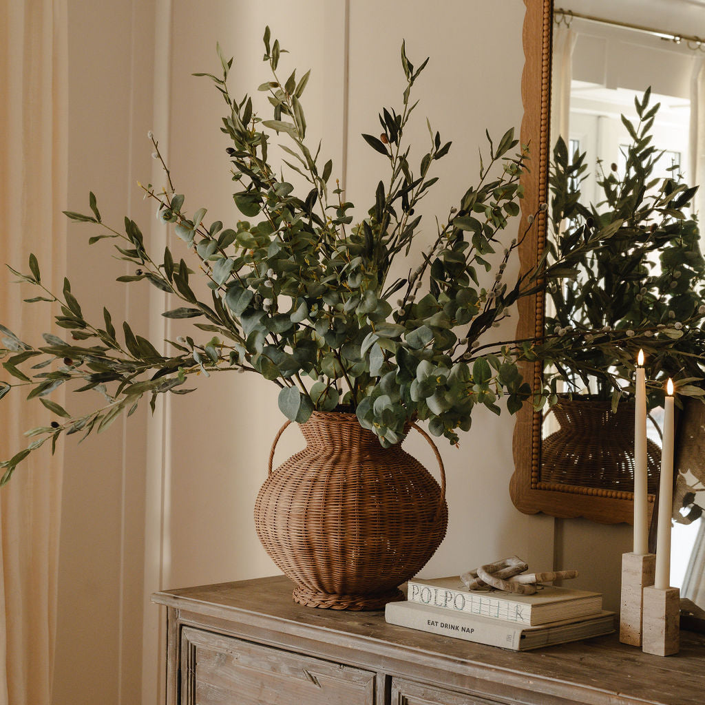 A woven wicker vase with a plant arrangement on a wooden surface, possibly a dresser or table, in a room with a mirror and chair.